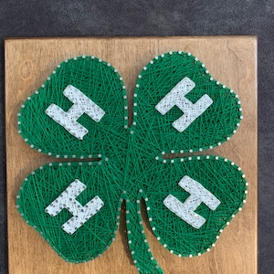May include: A wooden plaque featuring a four-leaf clover design made with green string. The clover has four white "H" letters, each within a leaf. The background is a light brown wood.