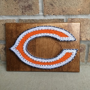 May include: A string art piece depicting the Chicago Bears logo. The logo is made of white, orange, and blue string wrapped around nails hammered into a wooden board.