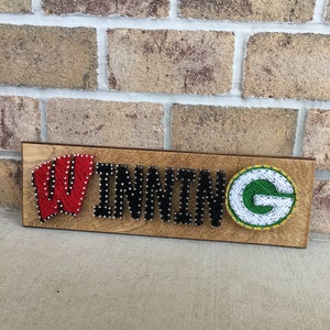 May include: Wooden sign with the word "WINNING" spelled out in black string. The "W" is red with white dots, and the "G" is green and yellow. The sign is on a brick background.