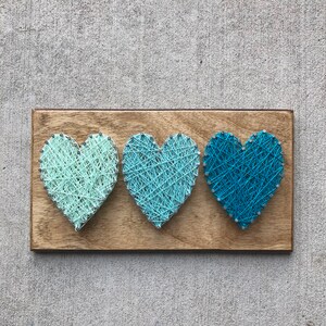 May include: A wooden plaque featuring three string art hearts in shades of blue and green. The hearts are arranged horizontally and are secured with small silver nails. The wooden base has a natural wood grain finish.