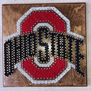 May include: String art of the Ohio State University logo. The logo is made of red, white, and black string wrapped around nails hammered into a wooden board.