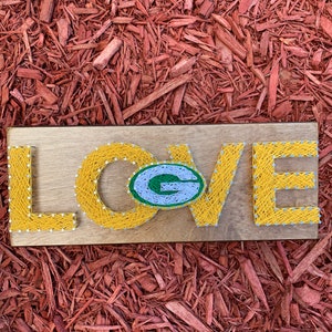 May include: A wooden sign with the word "LOVE" spelled out in yellow string. The "O" is replaced with a green and white Green Bay Packers logo.