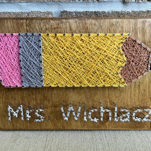 May include: A wooden sign with a string art pencil design in pink, gray, yellow, brown, and black. The sign also has the text "Mrs. Wichlacz" in white string art.