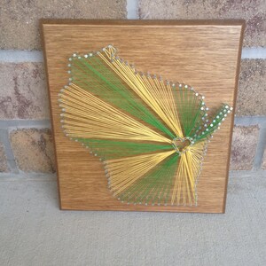 May include: A wooden plaque with a string art design of the state of Wisconsin. The string art is made with yellow and green string and is in the shape of the state. There is a small heart in the center of the design.