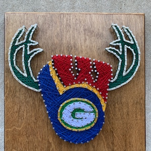 May include: String art piece on a wooden board featuring a stylized antler design. The design incorporates the Green Bay Packers logo with green, yellow, red, blue, and white string. The wooden board has a natural wood grain finish.