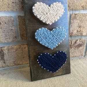 May include: Three string art hearts in shades of blue and white on a dark wood background. The hearts are arranged vertically, with the largest heart at the bottom and the smallest heart at the top.