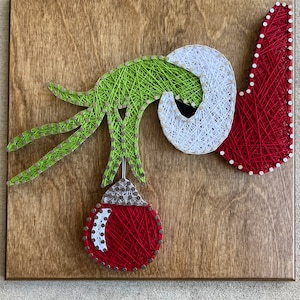 May include: Green, white, and red string art depicting the Grinch's hand reaching for a Christmas ornament.