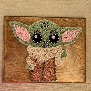May include: A string art piece depicting a green character with large eyes and pink ears. The character is made of string wrapped around nails hammered into a wooden board.