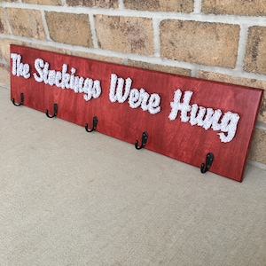 May include: A red wooden sign with the words "The Stockings Were Hung" in white string art. The sign has five black metal hooks.