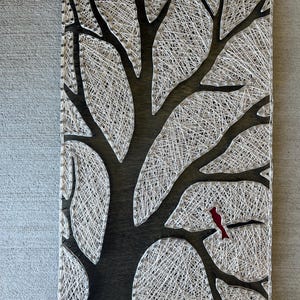 May include: A rectangular wooden wall art piece featuring a tree design created with white string and dark wood. The tree's silhouette is filled with string, and a small red bird is perched on a branch. The artwork is mounted on a gray surface.