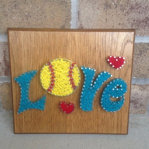 May include: A wooden sign with the word "LOVE" spelled out in blue yarn wrapped around nails. A yellow softball with red stitching is in the center of the sign. Two small red hearts are on either side of the softball.