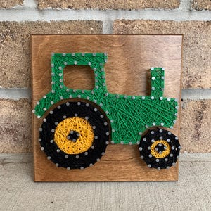 May include: Green tractor string art on a wooden background. The tractor has black wheels with yellow centers.