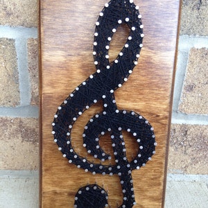 May include: A wooden plaque with a stained finish features a black string art treble clef design. The musical symbol is created with black thread wrapped around small silver nails, creating a striking contrast against the wood.
