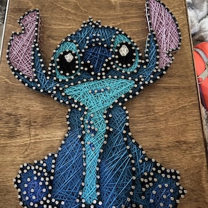 May include: A handmade string art piece featuring the character Stitch from Lilo & Stitch. The artwork uses blue, teal, and purple thread on a wooden board. The character's eyes are embellished with sparkling accents.