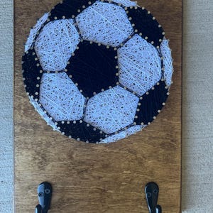 May include: A wooden wall hanging with a black and white string art soccer ball design. The hanging has two black metal hooks on the bottom.