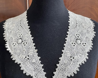 May include: An off-white, vintage lace collar with a scalloped edge. The collar is displayed on a black mannequin, showcasing its intricate floral and geometric patterns. The collar is a delicate piece of antique clothing.
