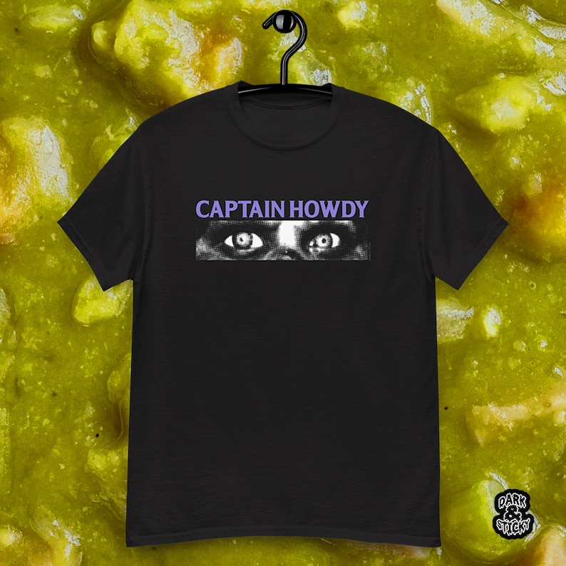 May include: Black t-shirt with a white image of a pair of eyes and the text "CAPTAIN HOWDY" in purple.