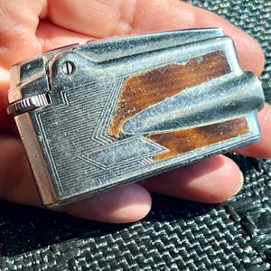 May include: A vintage silver-toned lighter with a rectangular shape and a decorative geometric design. The lighter features a brown, possibly wooden, accent on the side. The lighter is held in a hand.