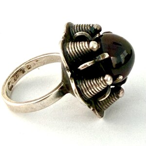 May include: A silver ring with a dark, round gemstone. The ring features a unique design with coiled metal accents surrounding the stone. The band is simple and polished, with markings visible on the inside.