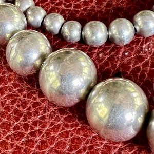May include: A close-up of a silver beaded necklace with large, round beads. The beads are smooth and shiny, and they are arranged in a single strand. The necklace is lying on a brown leather surface.