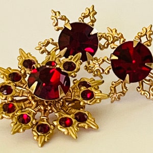May include: A pair of gold tone brooches with red rhinestone accents. The brooches are in the shape of a starburst with intricate details.