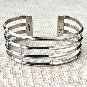May include: A silver cuff bracelet with a layered design. The bracelet has four horizontal bars and two vertical bars at the ends. The bracelet is made of a shiny metal and has a simple, elegant design.