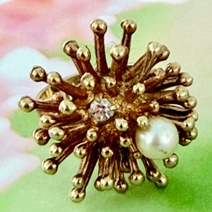 May include: A gold-toned ring with a spiky, starburst design. The ring features a central clear gemstone and a white pearl. The ring's design includes numerous small, round gold accents, creating a textured and eye-catching appearance.