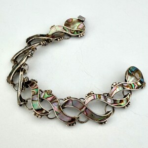 May include: A silver-toned bracelet with an intricate design. The bracelet features iridescent abalone shell inlays in a series of interconnected, flowing shapes. The clasp is a simple hook and eye closure. The overall design is elegant and eye-catching.