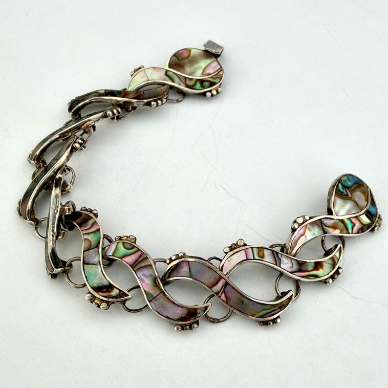 May include: A silver-toned bracelet with an intricate design. The bracelet features iridescent abalone shell inlays in a series of interconnected, flowing shapes. The clasp is a simple hook and eye closure. The overall design is elegant and eye-catching.