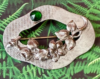 May include: An oval-shaped silver-toned brooch featuring a textured surface with a single round faceted emerald green stone at the top left and a raised relief design of leaves and a flower extending across the lower right section of the frame.
