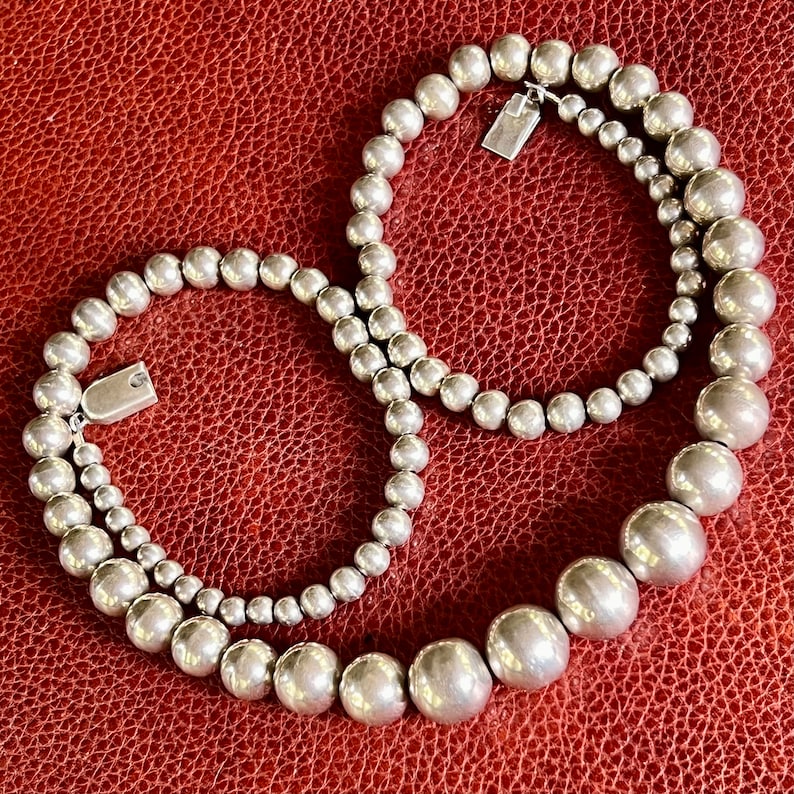 May include: A silver beaded necklace with two strands of varying sizes. The necklace is made of round beads and has a clasp closure.
