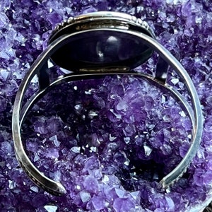 May include: A silver bracelet featuring a round, dark purple stone set in a bezel setting. The bracelet is open and has a hinged clasp.