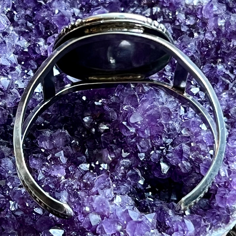 May include: A silver bracelet featuring a round, dark purple stone set in a bezel setting. The bracelet is open and has a hinged clasp.