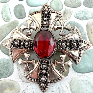 May include: A silver-toned metal brooch shaped like a cross, featuring a large, oval-cut, deep red faceted gemstone at its center. The cross is decorated with intricate floral metalwork patterns and four smaller cross shapes at the corners.