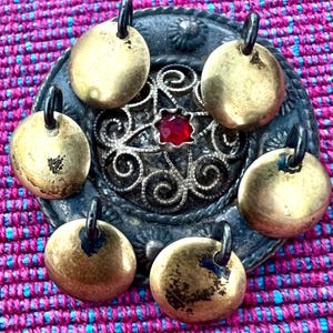 May include: A collection of antique brass pendants with black loops, arranged around a decorative silver-toned metal centerpiece with a red gemstone. The pendants have a worn, aged appearance. The background is a vibrant pink and blue patterned fabric.
