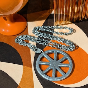 May include: A silver-toned metal necklace featuring a circular pendant with a spoked wheel design. The pendant is attached to a medium-weight link chain. The metal has a matte, slightly textured finish throughout the entire piece of jewelry.