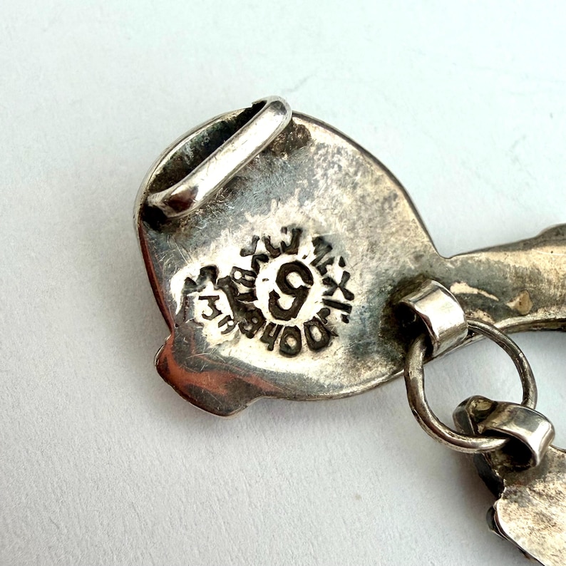 May include: Close-up of a silver-toned pendant with a detailed design. The pendant features a loop for hanging and a stamped marking with the number 9 and the word "Mexico". The metal has a slightly aged appearance.