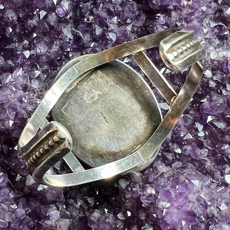 May include: A silver bracelet with a round, silver-toned face. The back of the face is stamped with the word "STERLING".