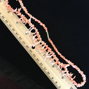 Op de afbeelding: Een roze koraal ketting met een dubbele rij kleine kralen en een enkele rij grotere koraaltakken. De ketting wordt gemeten met een liniaal, wat aangeeft dat deze ongeveer 25 cm lang is.