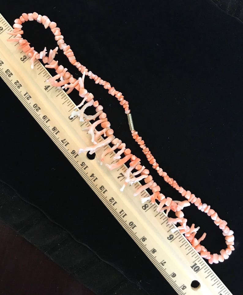 Op de afbeelding: Een roze koraal ketting met een dubbele rij kleine kralen en een enkele rij grotere koraaltakken. De ketting wordt gemeten met een liniaal, wat aangeeft dat deze ongeveer 25 cm lang is.