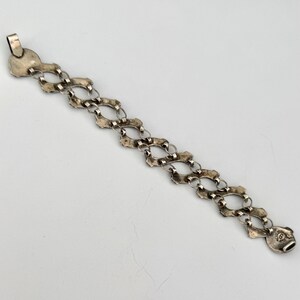 May include: A silver-toned bracelet with an ornate, open-link design. The bracelet features a series of interconnected, oval-shaped links with decorative edges. The clasp is a circular design with a decorative pattern.