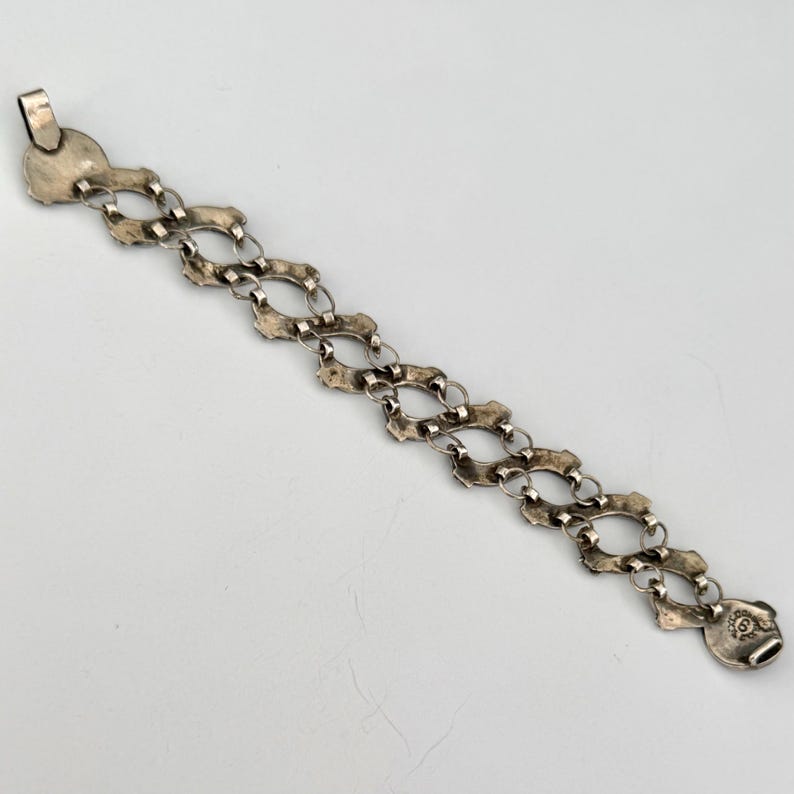 May include: A silver-toned bracelet with an ornate, open-link design. The bracelet features a series of interconnected, oval-shaped links with decorative edges. The clasp is a circular design with a decorative pattern.