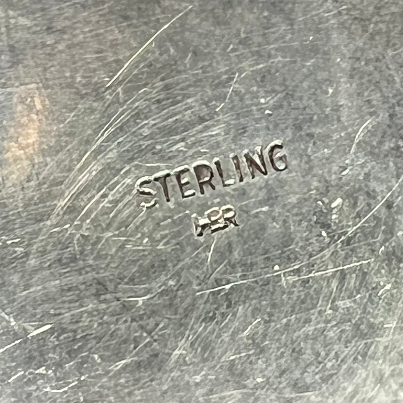 May include: Close-up of a silver surface with the word "STERLING" and the letters "HSR" stamped on it.