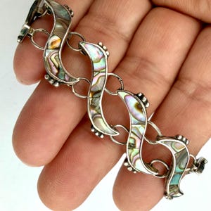 May include: A silver bracelet with iridescent abalone shell inlays. The bracelet features a series of curved, linked segments, each with a pearlescent, multi-colored surface. Small, decorative silver beads accent each segment.