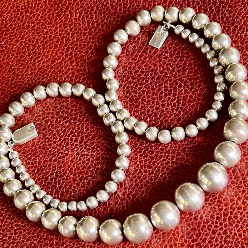 May include: Two silver necklaces with round beads. The necklace on top has smaller beads and the necklace on the bottom has larger beads.