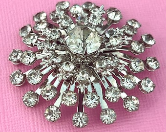 May include: A silver brooch featuring a large, clear central rhinestone encircled by smaller, clear rhinestones. The brooch has a starburst design with radiating lines, set against a pink background.