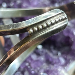 May include: Close-up of a silver bracelet with a decorative beaded edge. The bracelet is made of smooth, polished metal and has a simple, elegant design.
