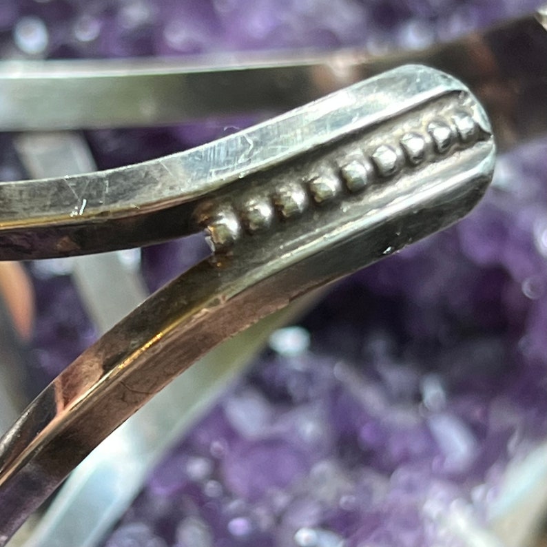 May include: Close-up of a silver bracelet with a decorative beaded edge. The bracelet is made of smooth, polished metal and has a simple, elegant design.