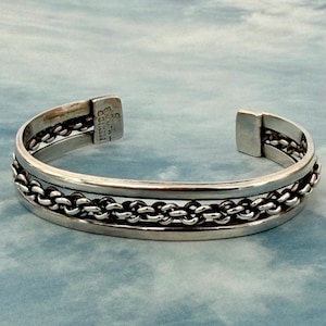 May include: A silver cuff bracelet with a chain link design. The bracelet has a rectangular clasp and is made of a shiny metal. The bracelet is on a blue and white background.