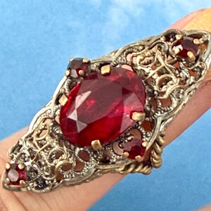 May include: An ornate ring with a large, faceted, oval-shaped red gemstone as the centerpiece. The ring has a filigree design with smaller red stones and gold accents. The ring is set against a blue background.